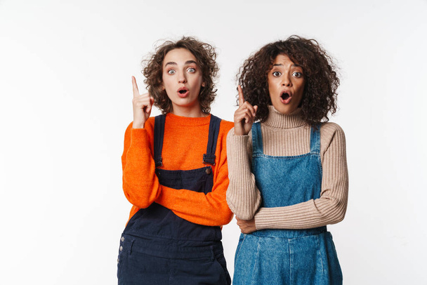 Portrait of shocked multinational women in overalls looking at camera and pointing fingers upward isolated over white background - 写真・画像