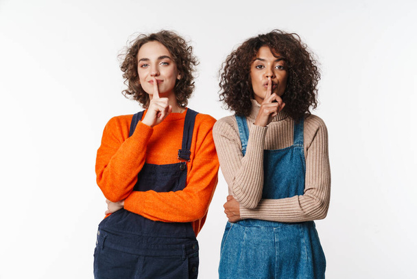 Portrait of curly multinational women in overalls gesturing silence sign at camera isolated over white background - Foto, Imagem