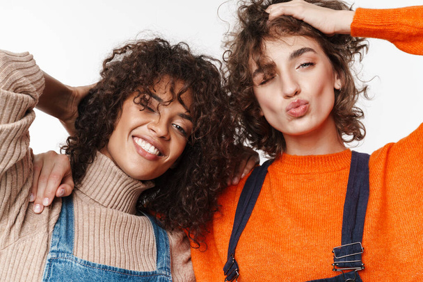 Image of a pretty happy smiling two multiracial girls friends in denim jumpsuits isolated over white wall background. - Foto, immagini