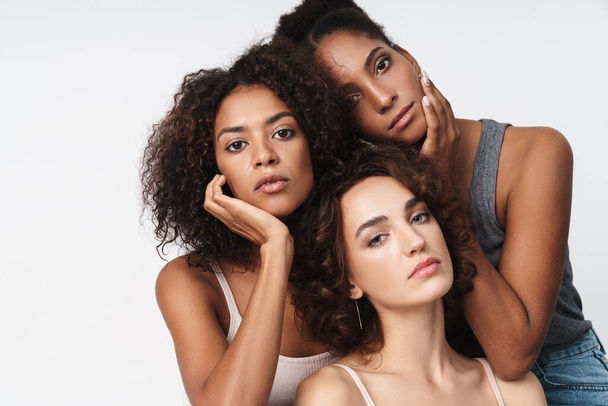 Portrait of three gorgeous multiethnic women standing together and looking at camera isolated over white background - Zdjęcie, obraz
