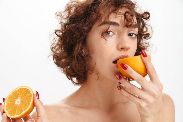 Close up beauty portrait of an attractive young topless woman with short curly brown hair isolated over white background, biting an orange - Fotoğraf, Görsel