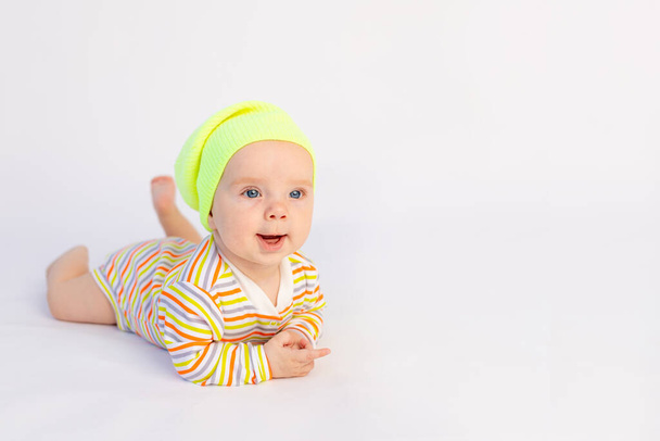 small smiling baby girl 6 months old lies on a white isolated background in a bright bodysuit and yellow hat, space for text - Photo, Image