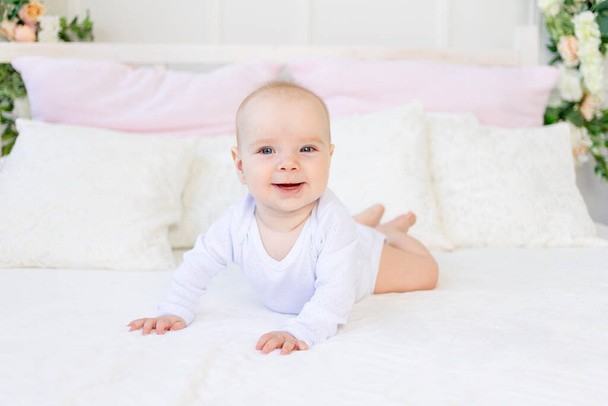 boldog kislány 6 hónapos, fehér ruhában fekszik a hasán egy fehér ágyon otthon. - Fotó, kép