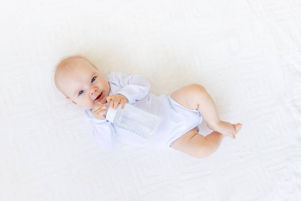 klein baby meisje 6 maanden oud in een wit pak liggend op haar rug op een wit bed thuis met een fles water, baby food concept - Foto, afbeelding