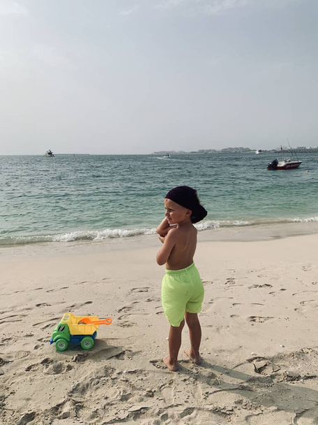 Песчаный морской пляж с малышом в ярких шортах и загорелой коже с пластиковым игрушечным самосвалом, семейный отдых, концепция отдыха - Фото, изображение