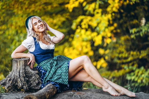 Oktoberfest, St. Patrick day, intenational beer day. Barefoot happy pretty blond girl in dirndl, traditional beer festival dress, sitting outdoors with blured colorful trees behind - Fotografie, Obrázek