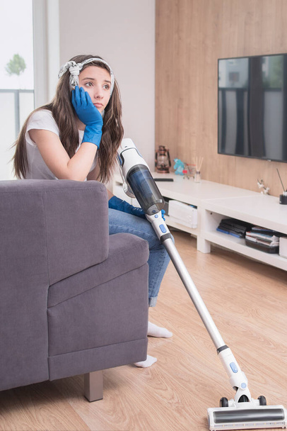 housekeeper .woman using vacuum cleaner in the living room.A young girl tired of cleaning the house is sitting on the sofa with a vacuum cleaner. - Zdjęcie, obraz
