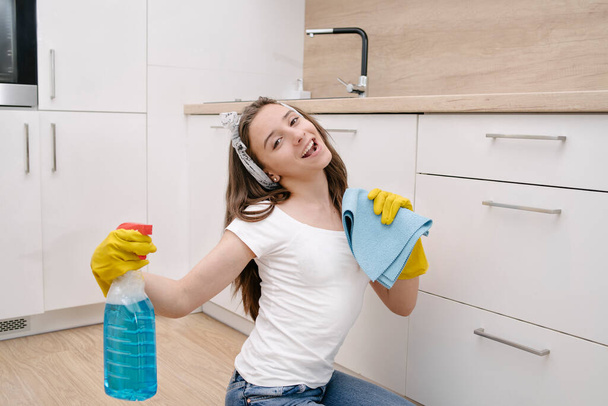 Ev temizliği servisi. Koruyucu eldivenli neşeli genç bir kız mutfağı yıkıyor ve şarkı söylüyor. Evi temizleyen, şarkı söyleyen mutlu bir kadın. Ev işleri, ev işleri. Evi temizleyen kız.. - Fotoğraf, Görsel