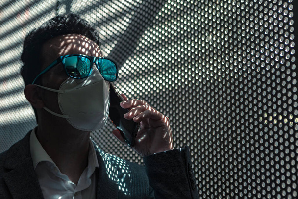 Low angle of unrecognizable male executive manager in formal wear and sunglasses with protective mask having phone call while standing near mesh wall with shadow - Foto, Imagen