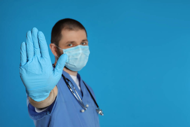 Doctor in protective mask showing stop gesture on light blue background, space for text. Prevent spreading of coronavirus - Foto, afbeelding