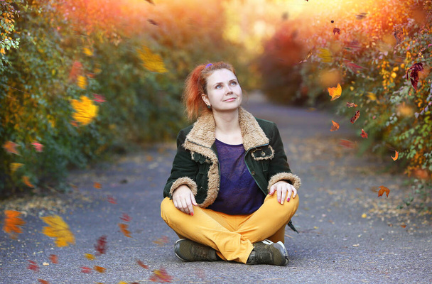 Happy young woman in lotus position with a smile sitting on the asphalt path in the autumn park. Leaf fall. Autumn concept. Copy space on the left - Photo, Image