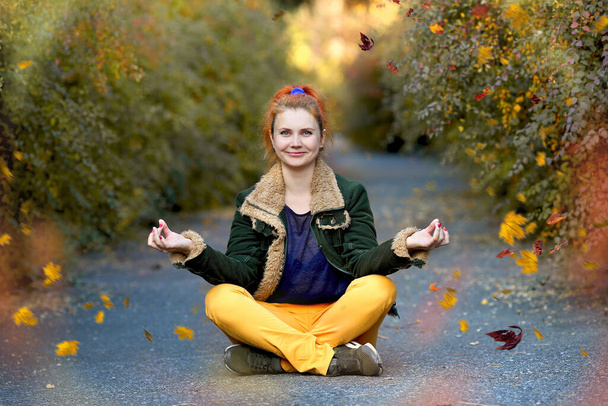 A young girl with a red tail smiling sits in a lotus position in an autumn park. The girl is dressed in yellow jeans, a green jacket.  Autumn women's clothing. Autumn concept - Photo, Image