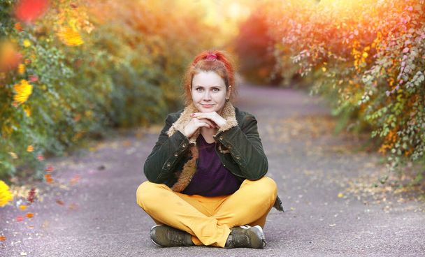 Happy young woman in lotus position with a smile sitting on the asphalt path in the autumn park. Leaf fall. Autumn concept. Copy space - Photo, Image