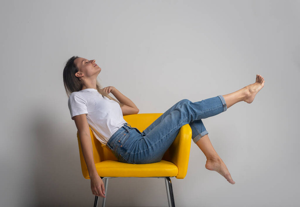 Beautiful young woman is sitting or lying on yellow chair with legs up and closing eyes with satisfaction and happiness, studio shot - Фото, изображение