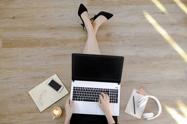 Top view of young woman wearing black dress sitting on wooden floor and working on her laptop. Cropped shot of female's feet. Close up, copy space, background, overhead. - Photo, image