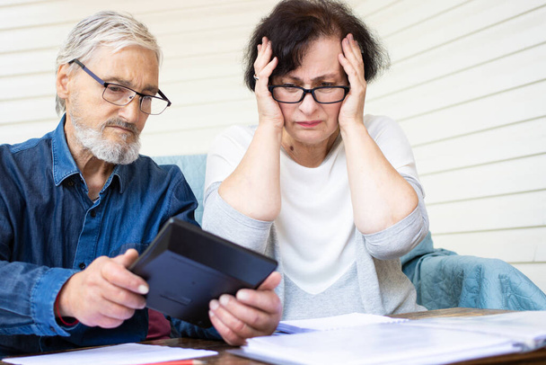 Verängstigtes und besorgtes älteres Ehepaar blickt auf Taschenrechner, zählt Bankkredite, berechnet Rechnungen, die die heimischen Finanzen verwalten. Sorgen wegen Konkurs oder Geldproblemen. Wenig Sparkonzept - Foto, Bild