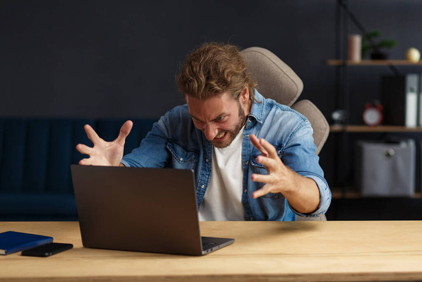 Angry young handsome curly man using laptop. Stressed freelancer lost data on broken not working laptop. Receiving bad news or email, spam message. Young people working with mobile devices - Foto, Imagen