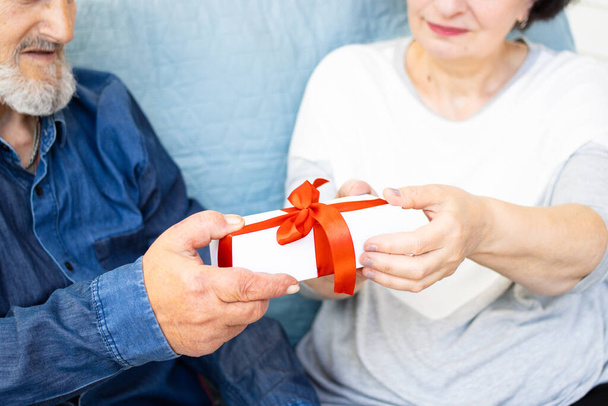 Nahaufnahme der Geschenkbox mit roter Schleife in den Händen eines älteren Ehepaares. Reifer Mann, der seine Frau überrascht und seine Liebe und Gefühle zum Ausdruck bringt. Feier und Gratulation zum Geburtstag, Jubiläum oder Weihnachten - Foto, Bild