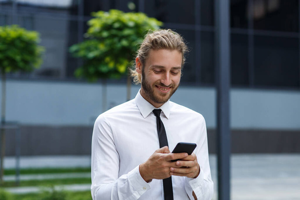 Portrait of a successful young businessman. A curly-haired man in a white shirt with a telephone against the background of a modern business center - Fotó, kép