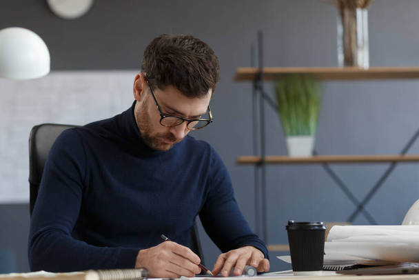 Architect working in office with blueprints.Engineer inspect architectural plan, sketching a construction project.Portrait of handsome bearded man sitting at workplace. Business construction concept - Фото, изображение