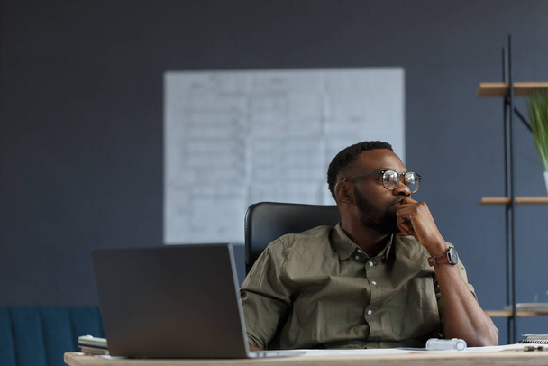 Afro-American architect working in office with laptop.Business portrait of black handsome bearded man wearing eyeglasses sitting at workplace.Confident businessman became successful.Business concept - Zdjęcie, obraz