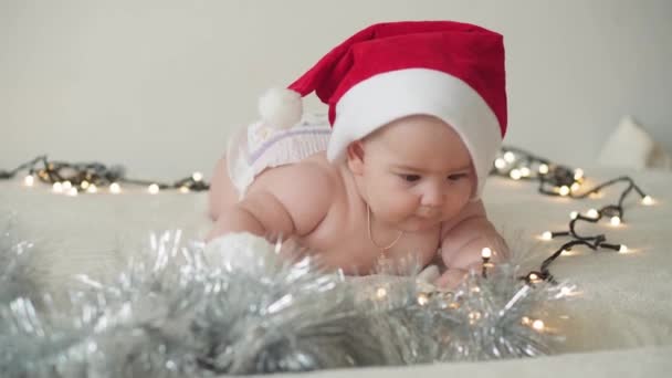 Feliz Navidad y feliz año nuevo, bebés, infancia, concepto de vacaciones - primer plano desnudo bebé recién nacido de 6 meses en sombrero de Santa Claus en su barriga se arrastra con decoraciones en el árbol de Navidad. - Imágenes, Vídeo