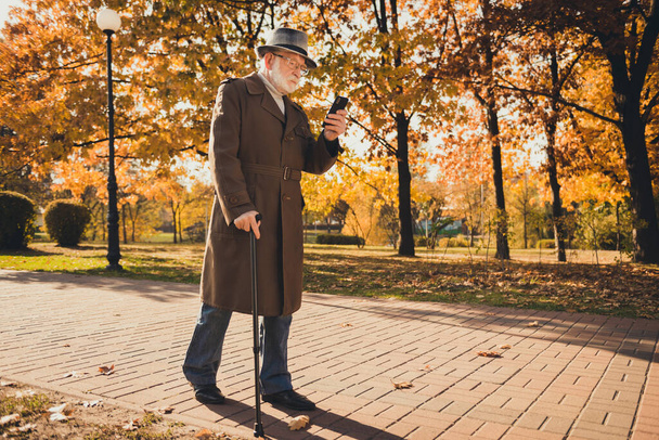 Full length profile photo of retired grey hair grandpa man walk desert park stick read email telephone sunny day use modern technology wear specs long coat headwear autumn colors outdoors - Foto, afbeelding
