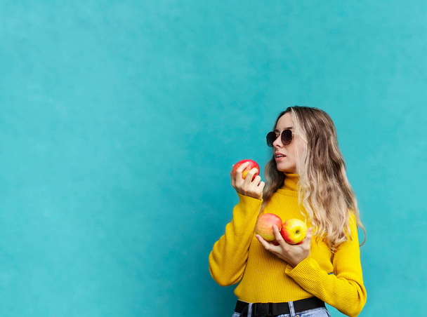 Blonde girl in yellow sweater with apples in her hands. Girl eating apples. Autumn harvest of fruits. Young woman on blue background in sunglasses - Zdjęcie, obraz