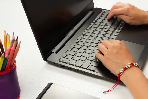 на белом деревянном столе лежат цветные карандаши, смартфон и ноутбук, на котором женские руки печатают текст - Фото, изображение