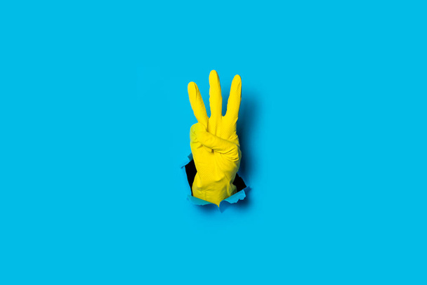 Hand in gelbem Handschuh mit drei Fingern auf hellblauem Hintergrund. - Foto, Bild
