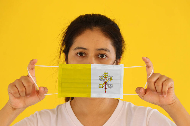 Eine Frau in weißem Hemd mit Vatikanstadt-Flagge auf Hygienemaske in der Hand und hochgezogenem Gesicht auf gelbem Hintergrund. Winzige Partikel oder Viren Corona oder Covid 19 Schutz. Konzept zur Bekämpfung von Krankheiten. - Foto, Bild