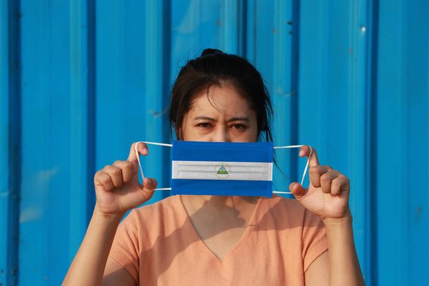 Une femme avec le drapeau du Nicaragua sur le masque hygiénique à la main et levé la face avant sur fond bleu. Protection minuscule contre les particules ou virus corona ou Covid 19. Concept de lutte contre la maladie. - Photo, image