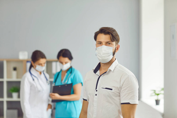 Man patient in medical protective mask standing over talking women doctors at background in clinic - Fotoğraf, Görsel