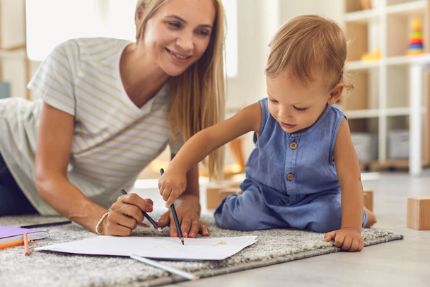 Adorable toddler scribbling on paper with pencil while sitting on floor with his young nanny - Photo, image