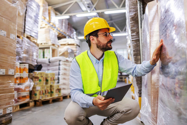 Young attractive bearded businessman in vest and helmet on head crouching and using tablet form checking on goods for shipment. Warehouse interior. - Foto, immagini