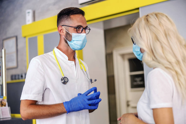 Médico joven con guantes de goma, mascarilla en uniforme estéril de pie en el hospital y llevar a un paciente acerca de los resultados de la prueba del virus corona. - Foto, imagen