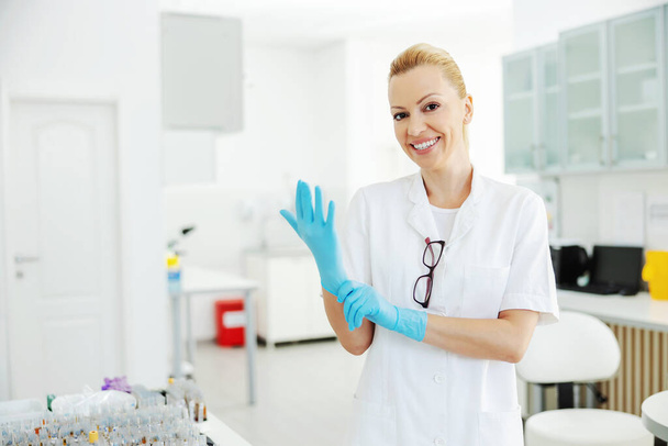 Middle aged attractive lab assistant in sterile white uniform standing in laboratory and putting on sterile rubber gloves and preparing herself to do research. - Foto, Bild