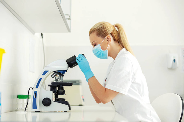 Bellissimo assistente di laboratorio con guanti di gomma e maschera facciale seduto in laboratorio e cercando attraverso il microscopio. - Foto, immagini