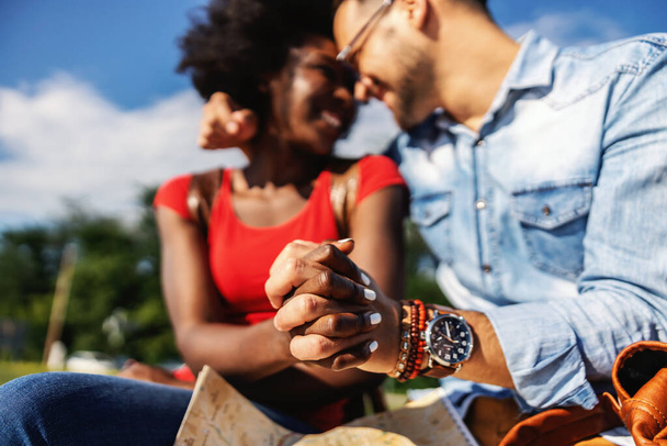 Nahaufnahme eines jungen lächelnden multikulturellen Hipster-Paares, das Händchen hält und an einem schönen sonnigen Tag im Freien sitzt. - Foto, Bild