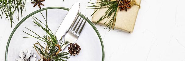 Kerst of Nieuwjaar tafelschikking. Feestelijke bestek, keramische borden met verse dennentak, kegels, nul afval geschenken en ster anijs. Witte klei achtergrond, banner formaat - Foto, afbeelding