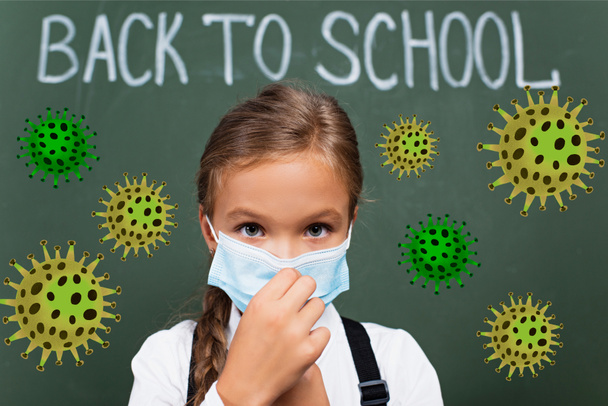 selective focus of schoolgirl touching medical mask near back to school lettering on chalkboard and virus illustration  - 写真・画像