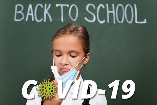 selective focus of displeased schoolgirl touching protective mask near chalkboard and covid-19 lettering in classroom  - Photo, Image