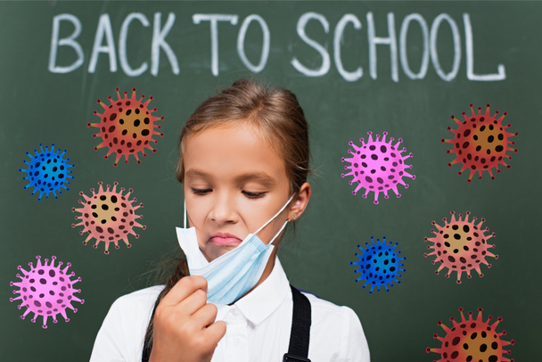 selective focus of displeased schoolgirl touching protective mask near chalkboard with back to school lettering and virus illustration  - Photo, Image