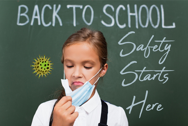 επιλεκτική εστίαση δυσαρεστημένη μαθήτρια αγγίζοντας προστατευτική μάσκα κοντά στο chalkboard με ασφάλεια ξεκινά εδώ γράμματα στην τάξη  - Φωτογραφία, εικόνα