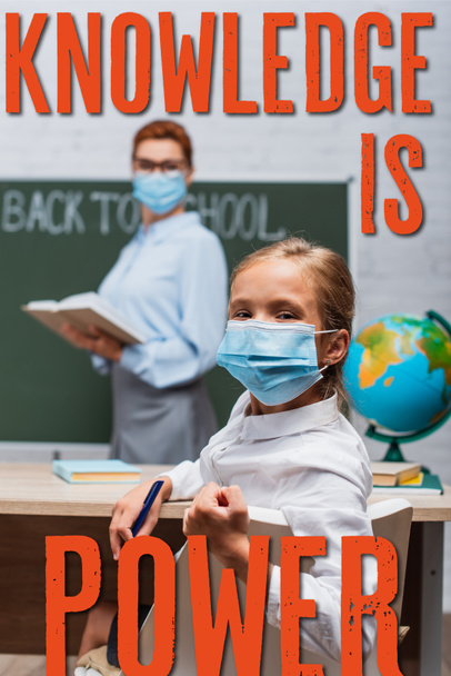 Selektiver Fokus der Schülerin in medizinischer Maske, die in die Kamera blickt, während der Lehrer neben der Tafel steht und Wissen Machtschrift ist  - Foto, Bild