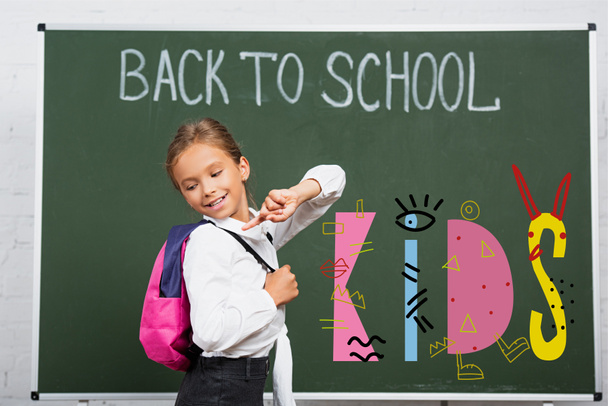 schoolgirl pointing with finger at backpack near chalkboard with back to school and kids lettering in classroom  - Photo, Image