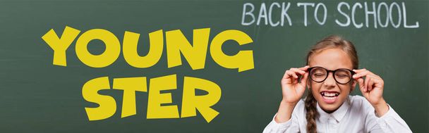 horizontal image of schoolgirl with closed eyes touching eyeglasses near chalkboard with back to school and youngster lettering in classroom  - Photo, Image