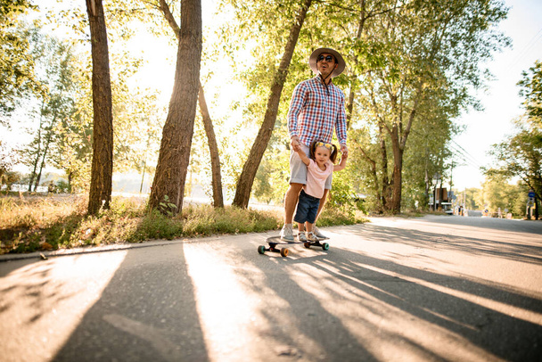 view on man who together with his little daughter riding skateboard on the road in summer park - Fotografie, Obrázek
