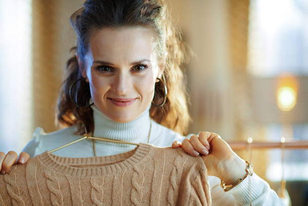 smiling trendy 40 years old woman in white sweater and skirt in the modern living room in sunny winter day holding sweaters on copper hanger. - Zdjęcie, obraz