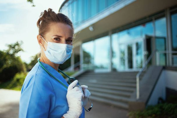 coronavirus pandemic. Portrait of confident modern physician woman in uniform with stethoscope and medical mask outside near clinic. - Фото, зображення